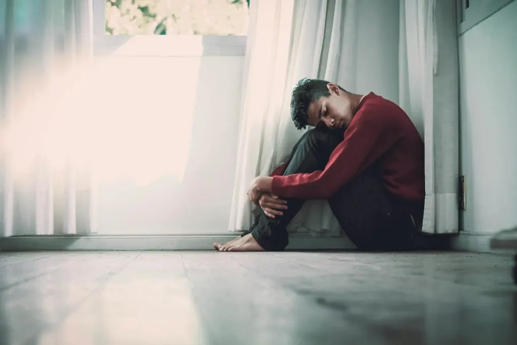 A grieving man sitting alone on the floor beside a window